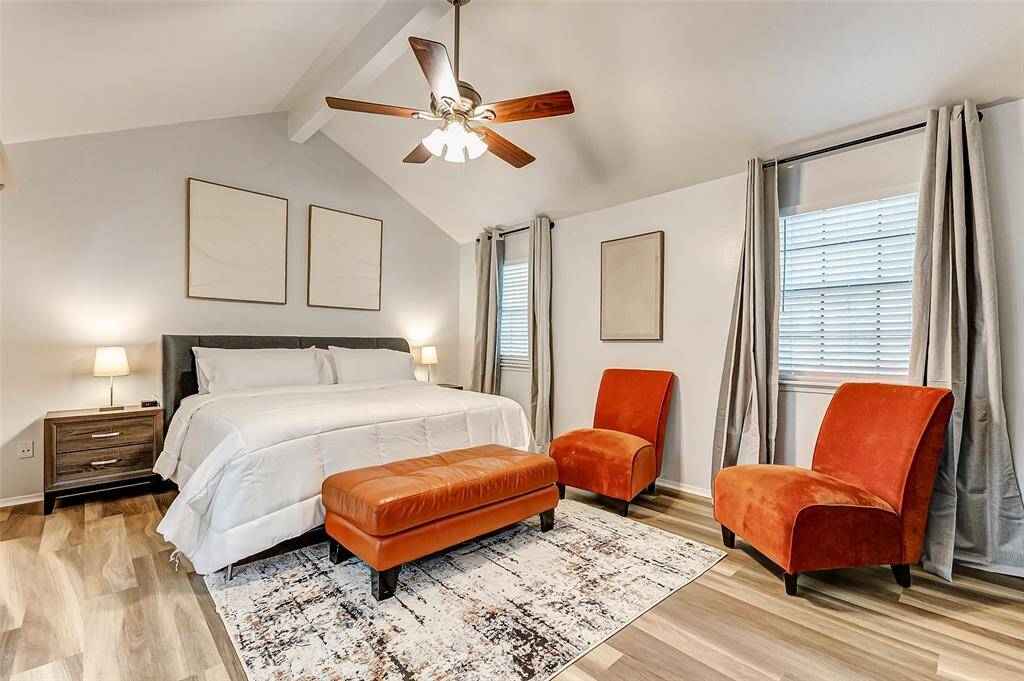This bedroom features a cozy and modern design with a vaulted ceiling, a ceiling fan, and natural light from two windows. It includes a comfortable bed, orange accent chairs, a patterned area rug, and neutral decor, creating a warm and inviting atmosphere.