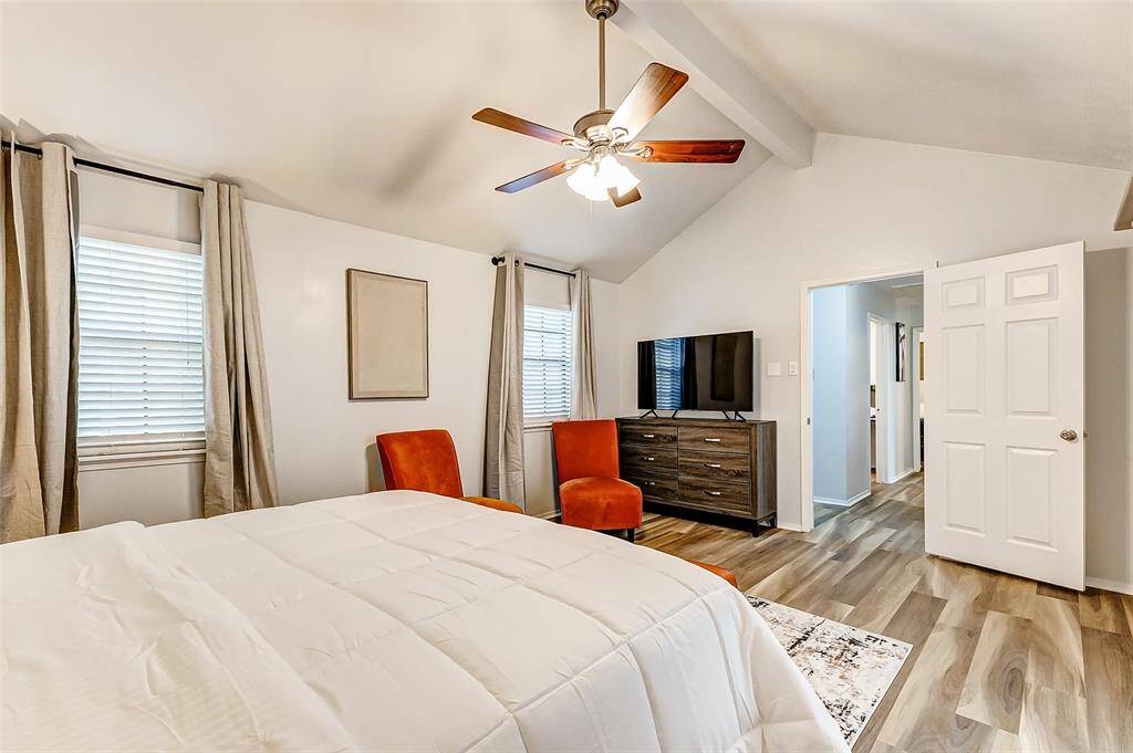 Bright and airy bedroom featuring a vaulted ceiling with a ceiling fan, large windows with curtains, and quality  flooring. It includes a cozy bed, a dresser with a TV, and two accent chairs, creating a comfortable and inviting space.