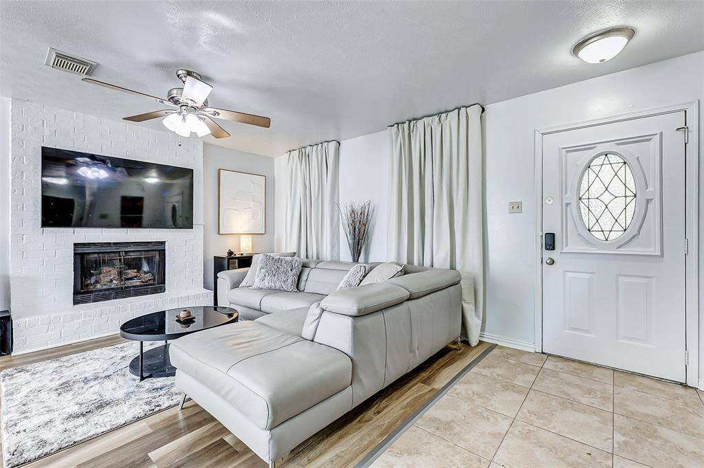 This cozy living room features a modern aesthetic with a white brick fireplace, mounted TV, and stylish ceiling fan. It includes a comfortable L-shaped sofa, a sleek black coffee table, and is accented by elegant curtains and a decorative front door. The space combines quality vinyl and tile flooring for a warm, inviting feel.