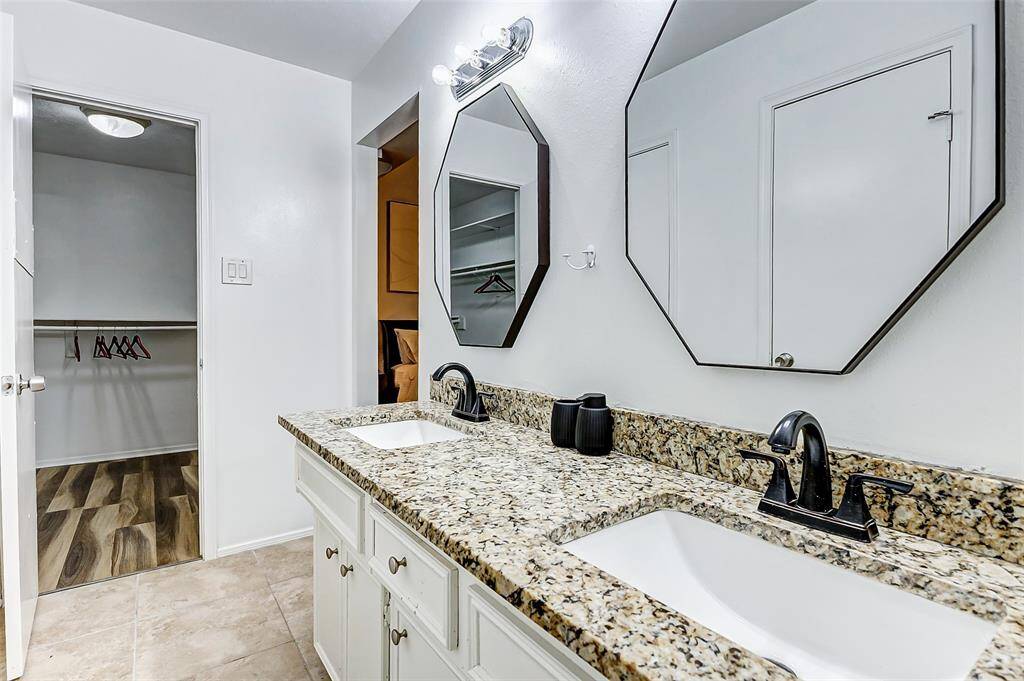 This is a modern bathroom featuring a dual-sink vanity with granite countertops and stylish black fixtures. It has geometric mirrors and leads into a spacious walk-in closet with stylish flooring.
