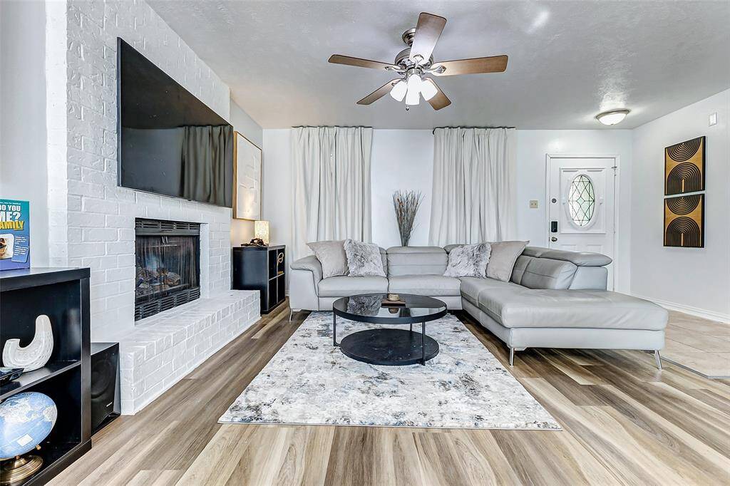 This inviting living room features a modern design with a white brick fireplace, mounted TV, and sleek ceiling fan. It has a spacious gray sectional sofa and a round black coffee table on a patterned rug, with light flooring enhancing the bright atmosphere.