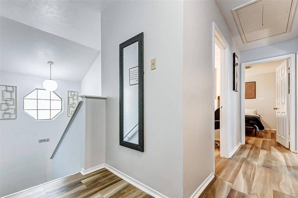 This photo features a bright, modern hallway with wood-look flooring, leading to bedrooms. A large window and stylish light fixture illuminate the space, complemented by a wall mirror and tasteful decor.