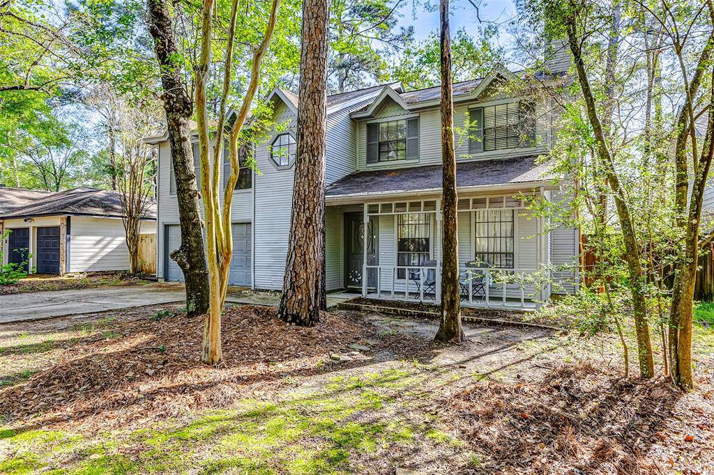 This charming two-story home features a classic facade with a covered front porch, nestled among mature trees for a serene, natural setting. It includes a double garage and a cozy outdoor seating area, offering both convenience and tranquility.