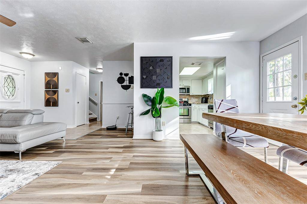This photo showcases a bright, open-concept living area with modern decor. It features stylish flooring, a sleek gray sectional, and a cozy dining space with a wooden table. The kitchen, visible in the background, has white cabinetry and stainless steel appliances, creating a contemporary and inviting atmosphere.