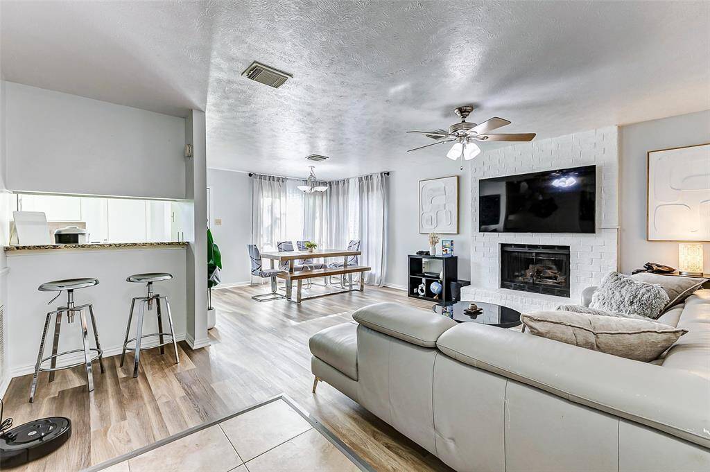 This inviting living space features an open layout with a cozy fireplace and a modern ceiling fan. The room is brightened by ample natural light from large windows, and it seamlessly flows into a dining area. A sleek bar counter with stools offers additional seating, and the light  flooring complements the contemporary decor.