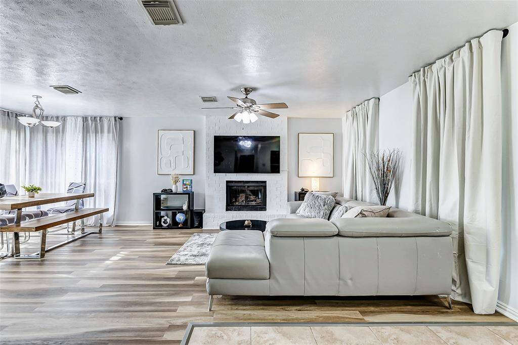 This spacious living room features a modern design with a cozy sectional sofa, a white brick fireplace, and a mounted TV. The open layout connects to a dining area with ample natural light from large windows, complemented by light color vinyl flooring and neutral decor.