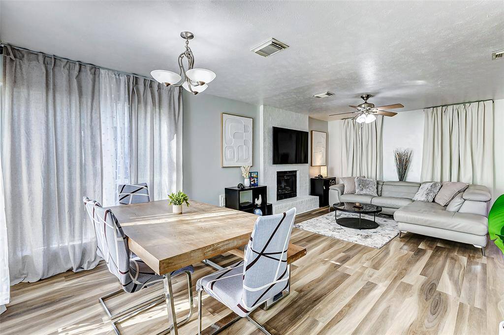 This inviting living and dining area features modern  flooring, ample natural light from large windows with sheer curtains, and a cozy fireplace. The space is tastefully decorated with a neutral color palette, a comfortable sectional sofa, and stylish lighting fixtures, providing a warm and welcoming atmosphere.