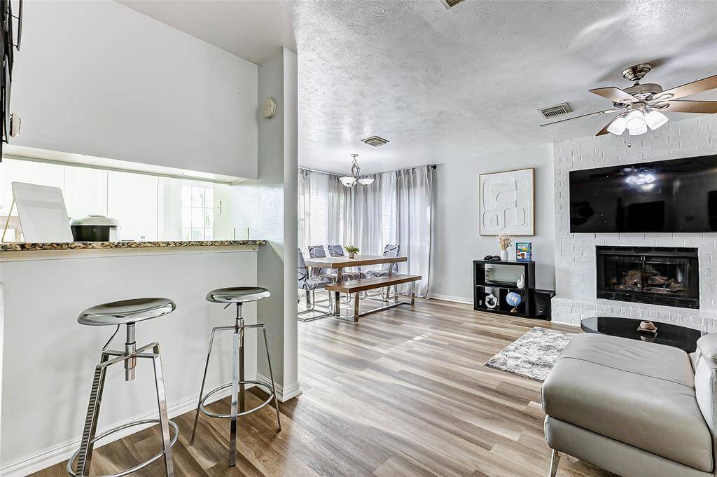 This inviting living space features an open layout with a cozy fireplace, modern ceiling fans, and natural light from large windows. The adjacent dining area offers a relaxed atmosphere, and the kitchen includes a convenient breakfast bar with sleek stools. Perfect for comfortable living and entertaining.