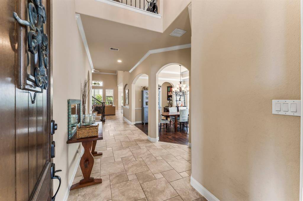 Step into sophisticated style with this stunning two-story foyer, where elegant upgrades like crown molding and two art niches add to the refined craftsmanship throughout. The grand Foyer welcomes you with warm Versailles-pattern tile flooring, a stately iron chandelier, convenient coat closet, and several areas for an entry table or bench. From the moment you enter, you're greeted with breathtaking views extending through the living room to the expansive backyard and welcoming pool beyond.