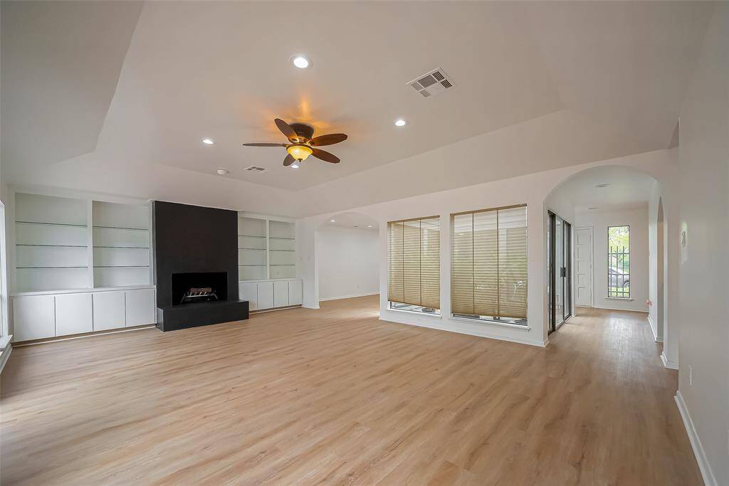 This spacious living area features a modern design with light vinyl flooring, a central black fireplace, built-in shelving, and a ceiling fan. Large windows with blinds allow natural light to fill the space, complemented by recessed lighting. An arched entryway leads to additional rooms, enhancing the open feel.