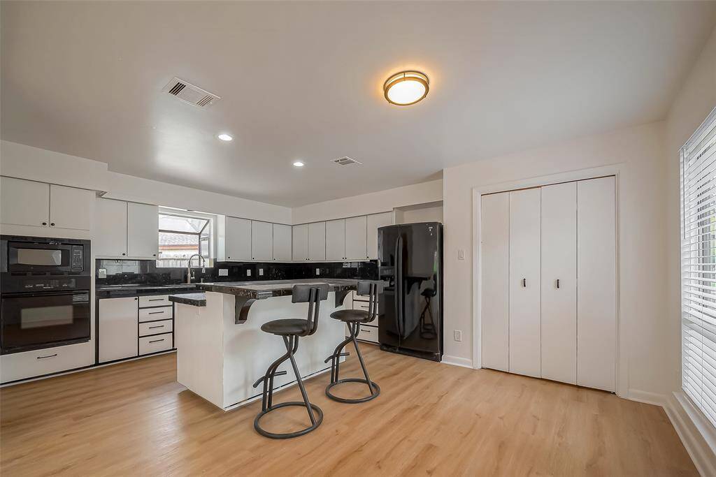 This kitchen features a modern design with white cabinetry, black countertops, and a center island with bar seating. It includes built-in appliances, a black refrigerator, and ample natural light from a large window. The space is complemented by light flooring and a cozy dining area.