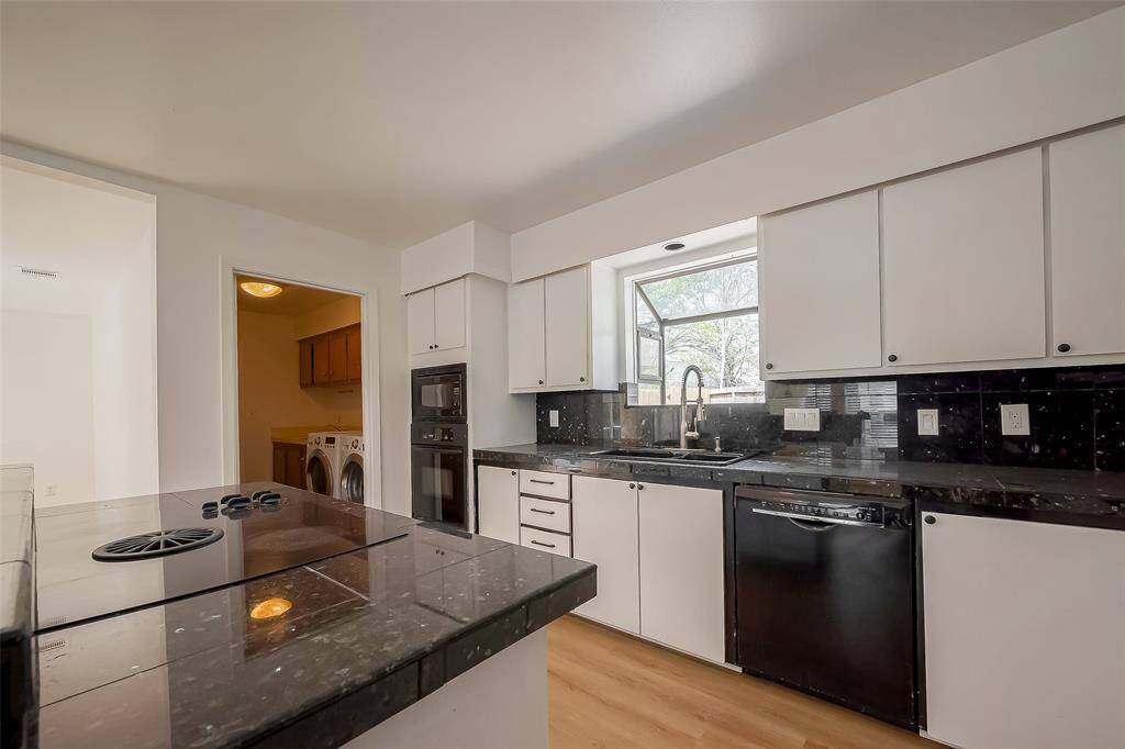 Modern kitchen with sleek black countertops and white cabinets, featuring a built-in oven, dishwasher, and electric cooktop. A large window above the sink provides natural light, and there's an adjacent laundry area with washer and dryer.