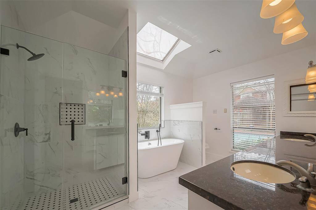 This bathroom features a spacious shower with glass doors, a freestanding tub under a skylight, dual sinks with granite countertops, and modern lighting. Natural light streams in through a large window, creating a bright and inviting space.