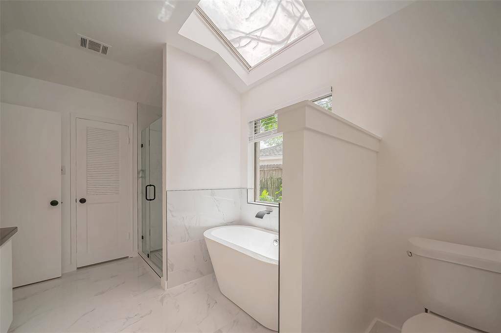 Bright and airy bathroom featuring a freestanding bathtub, glass-enclosed shower, and a skylight that enhances natural light. Clean, modern design with marble-style flooring and subtle finishes.
