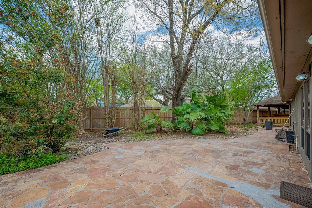 This photo showcases a spacious backyard with a stone patio, surrounded by lush greenery and mature trees. A wooden fence provides privacy, and a charming gazebo is visible in the corner, perfect for outdoor relaxation or entertaining.