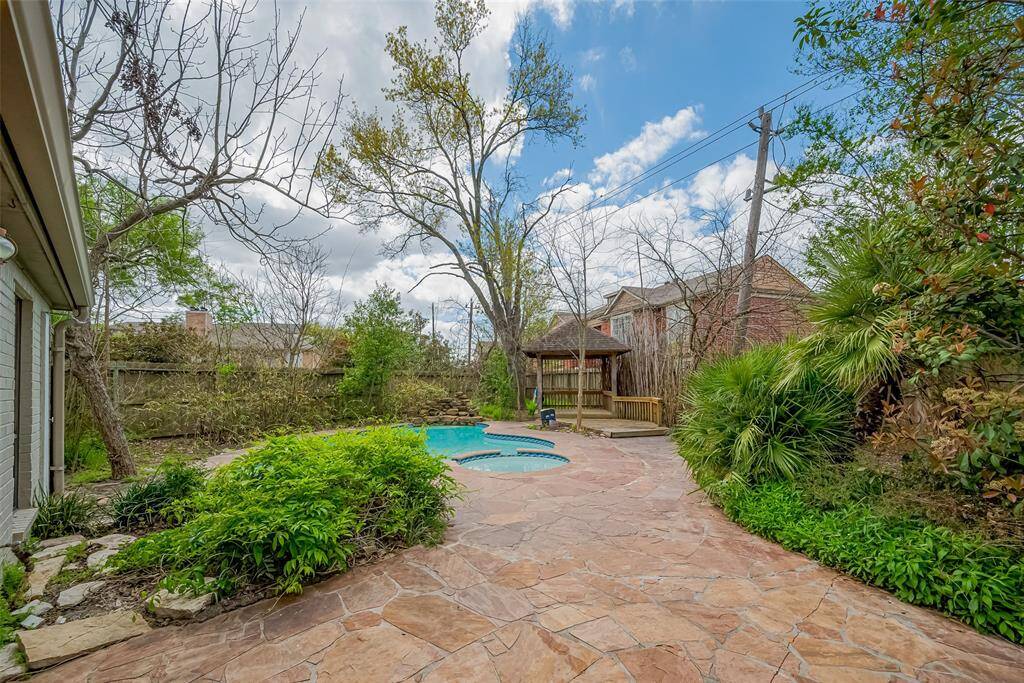 This backyard features a spacious stone patio, lush greenery, and a swimming pool. A wooden gazebo provides a cozy spot for relaxation, all surrounded by mature trees and privacy fencing.