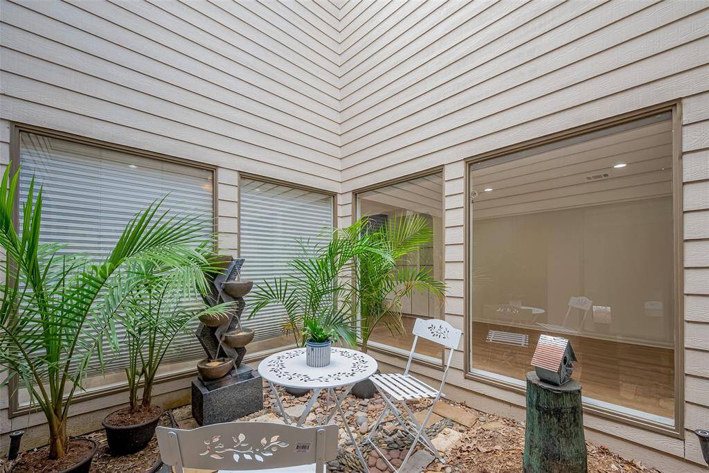 This photo showcases a cozy, private courtyard with a small table and chairs, surrounded by lush potted plants and a decorative water feature. Large windows provide ample natural light, enhancing the serene atmosphere.