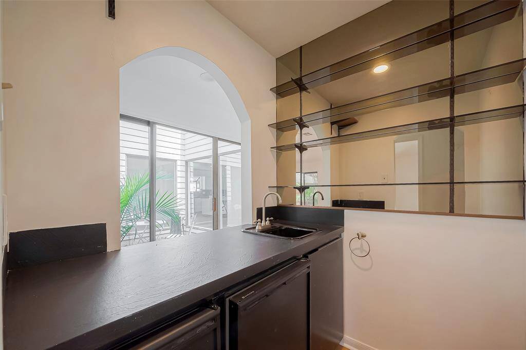 This photo showcases a cozy wet bar area with a sleek black countertop, built-in sink, and shelving. It opens to a bright, enclosed patio through an arched opening, adding a touch of modern elegance to the space.
