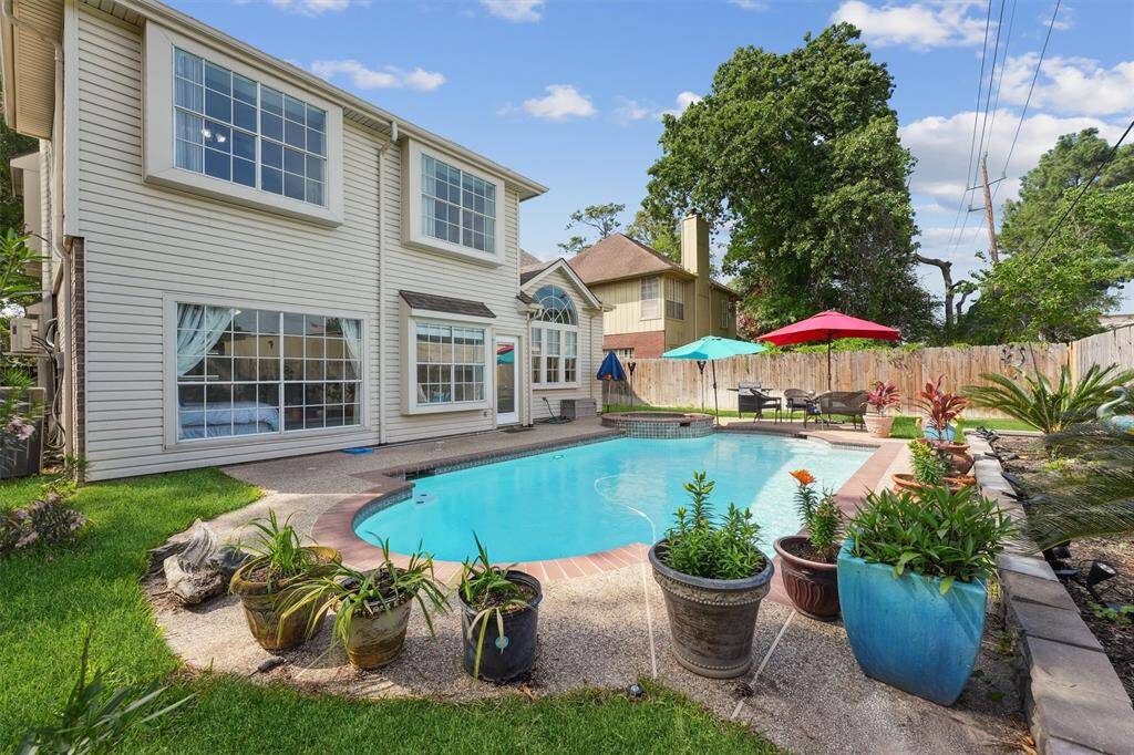 This inviting backyard oasis features a sparkling in-ground pool surrounded by lush greenery, vibrant potted plants, and a spacious patio perfect for relaxing or entertaining.
