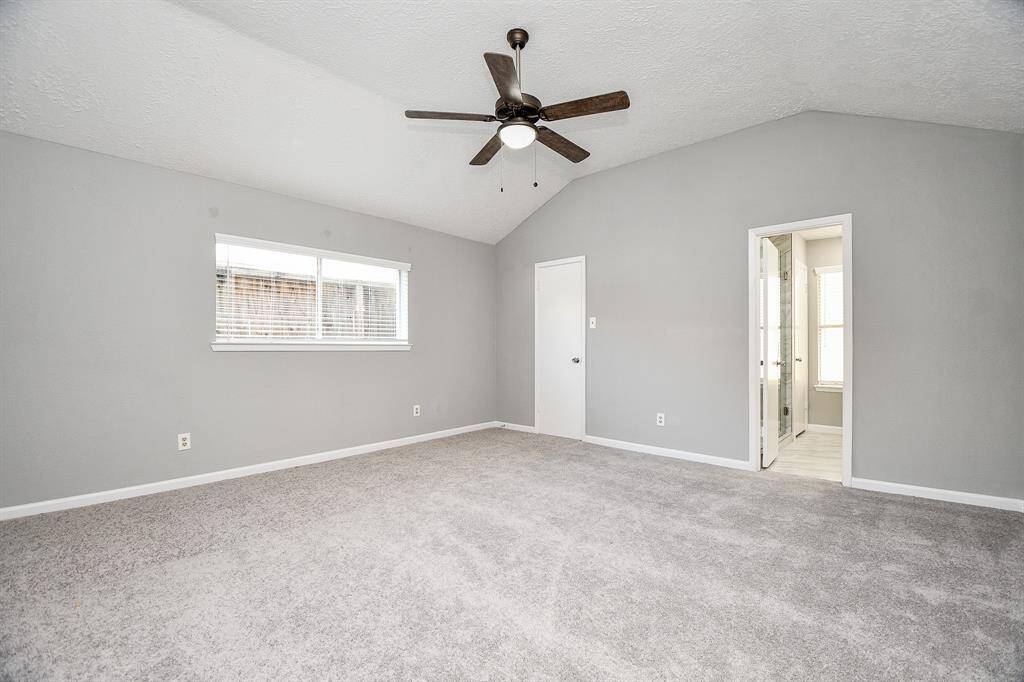This spacious master bedroom features soft gray walls, plush carpeting, and a ceiling fan. It offers ample natural light from a large window and includes an en-suite bathroom for convenience.