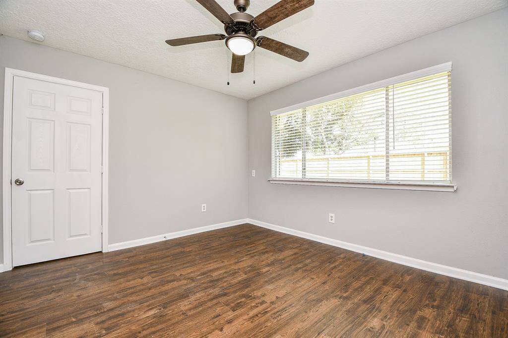 This room features neutral gray walls, a ceiling fan with light, and wood-style flooring. A large window provides ample natural light, and there's a white door for entry.