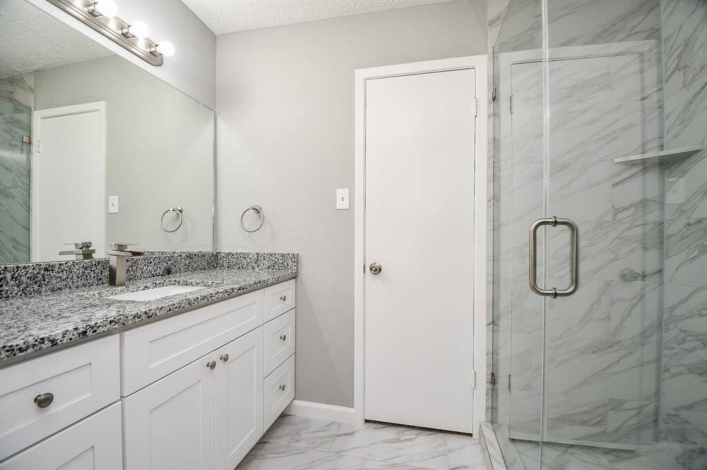 This bathroom features a modern design with a sleek glass shower, elegant gray and white marble tiles, a granite countertop with a large mirror, and ample cabinet space in white.
