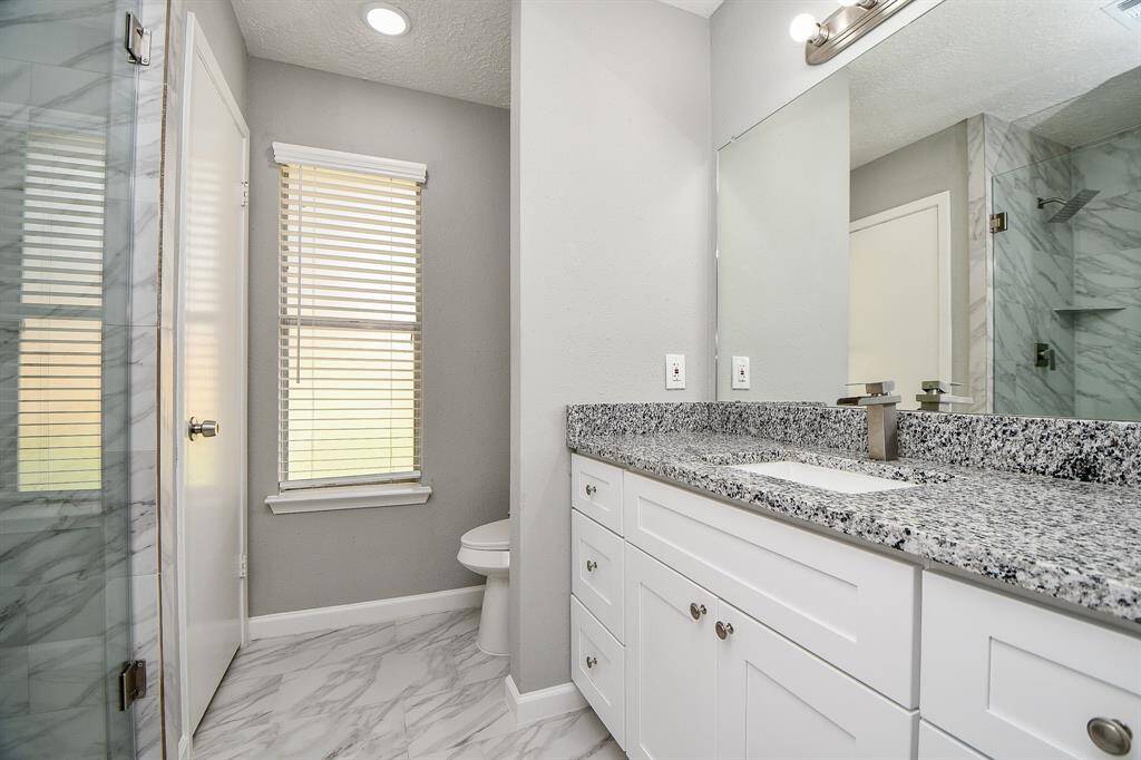 Modern bathroom featuring a sleek granite countertop, dual sinks, and a spacious glass-enclosed shower. The room is brightened by natural light from a window with blinds, complemented by a light neutral color palette.
