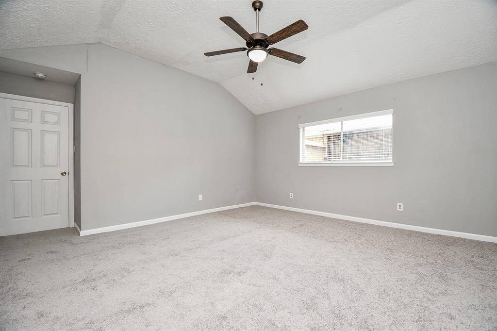 Bright and spacious bedroom with neutral gray walls and carpet, featuring a vaulted ceiling and modern ceiling fan. A large window allows natural light to fill the room.