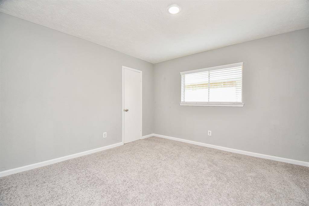 A bright, empty room with light gray walls and carpet, featuring a single window with blinds and a closed door. Ideal for a bedroom or office.