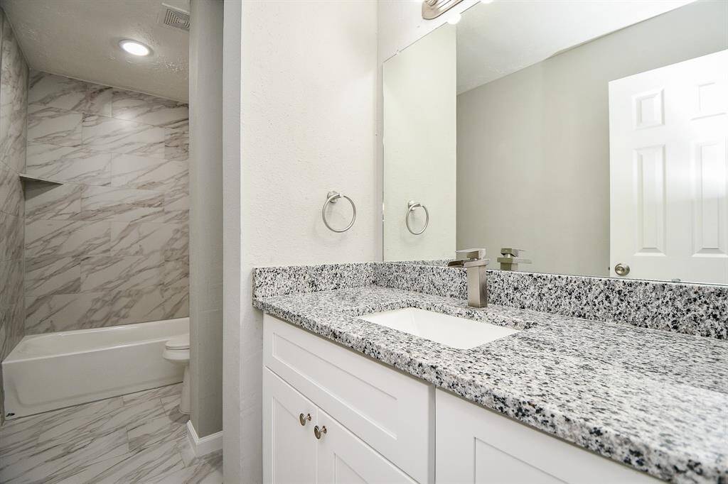 This bathroom features a modern design with a granite countertop and white cabinetry. The shower area is elegantly tiled with a marble-like finish, and recessed lighting provides a bright, clean ambiance.