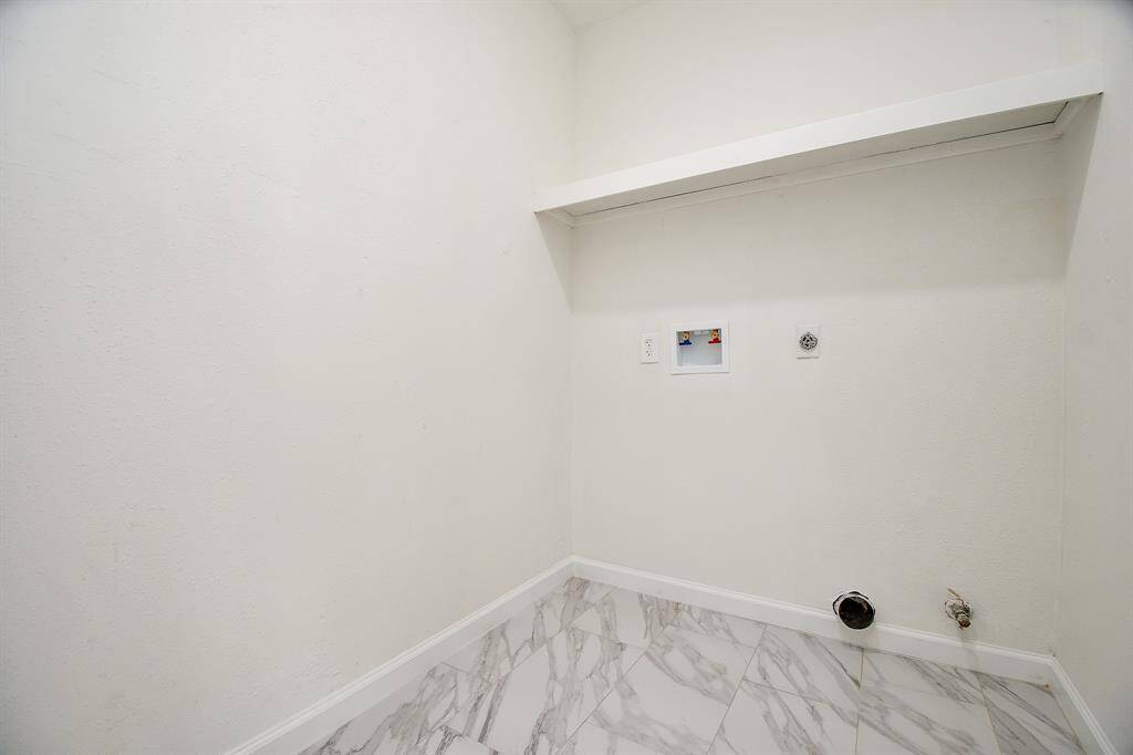 This photo shows a utility room with hookups for a washer and dryer. It features white walls, a shelf, and marble-patterned tile flooring.