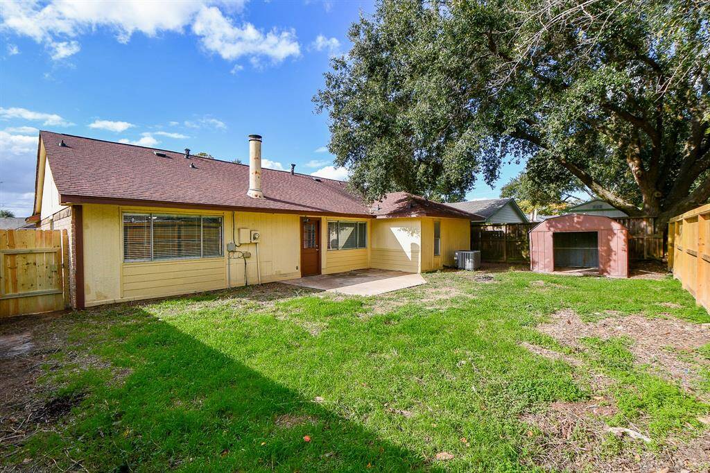 This photo showcases a charming backyard featuring a single-story home with a sloped roof, a spacious grassy area, and a large tree providing shade. There's a small shed in the corner and a wooden fence enclosing the yard, offering privacy.