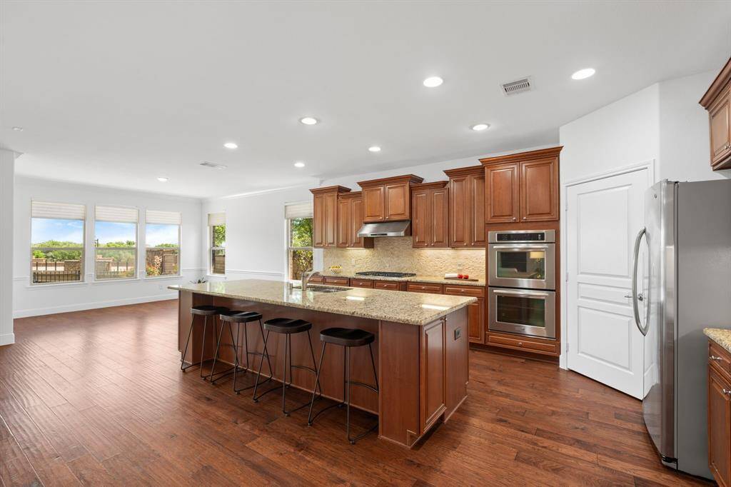 The kitchen features an oversized island with a convenient cabinet at the end, double ovens with convection capabilities, and a stunning stone backsplash. The beautiful wood flooring seamlessly flows throughout, tying the space together with both elegance and functionality.