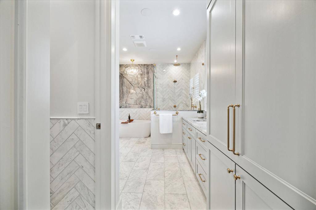 Spa-worthy primary bath with dramatic natural stone feature wall, soaking tub, herringbone marble tile, dual vanities, and large glass shower with rainfall fixture.