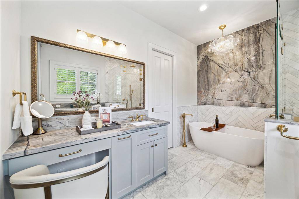 Gorgeous renovated primary bath with striking marble finishes, dual vanities, gold hardware, herringbone tile shower, and soaking tub set beneath a statement chandelier.