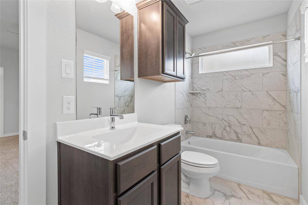 Secondary bath features tile flooring, bath/shower combo with tile surround, stained wood cabinets, beautiful light countertops, mirror, dark, sleek fixtures and modern finishes.