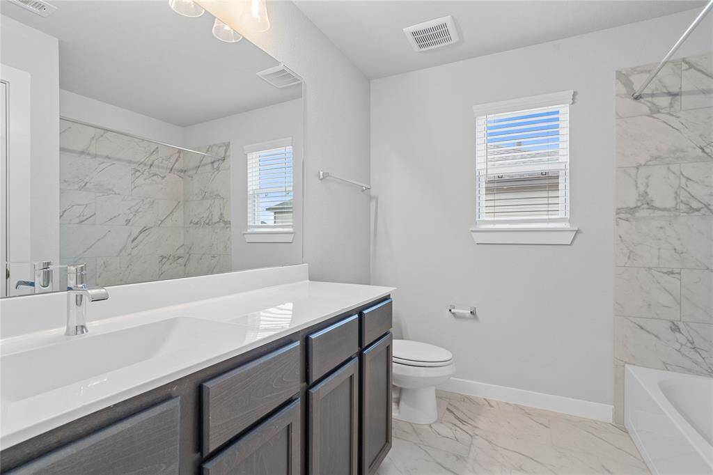 Secondary bath features tile flooring, bath/shower combo with tile surround, stained wood cabinets, beautiful light countertops, mirror, dark, sleek fixtures and modern finishes.