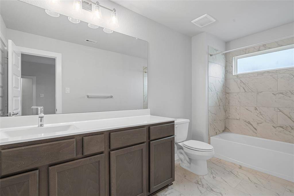 Secondary bath features tile flooring, bath/shower combo with tile surround, stained wood cabinets, beautiful light countertops, mirror, dark, sleek fixtures and modern finishes.
