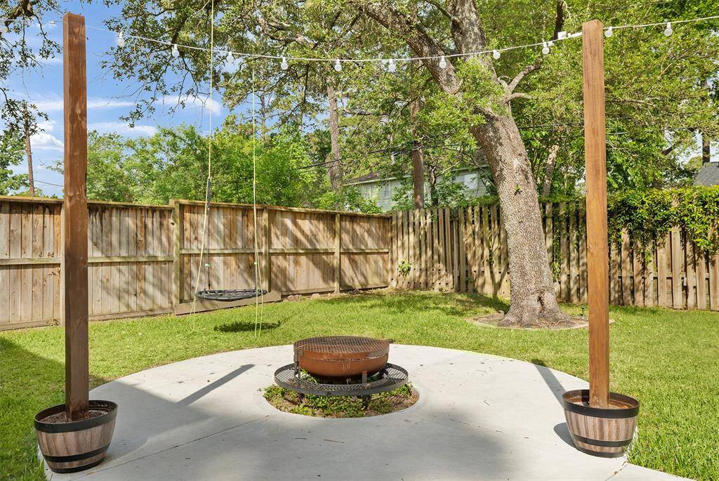 This backyard retreat invites you to unwind with a circular concrete patio centered around a cozy fire pit—perfect for evening gatherings under the stars. A swing hanging from a sturdy tree adds a playful touch, while string lights and a wooden fence create a warm, private atmosphere surrounded by nature.