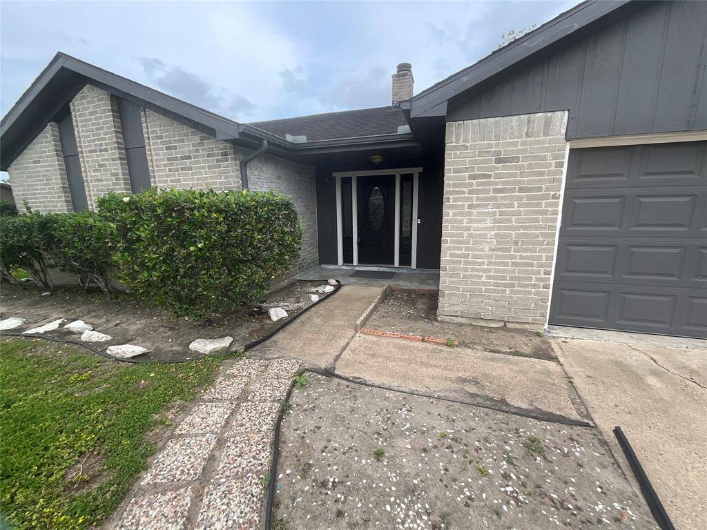 This photo shows the front entrance of a single-story home with gray brick exterior and a dark front door. A small pathway leads to the entrance, flanked by neatly trimmed bushes. There's an attached garage on the right side.