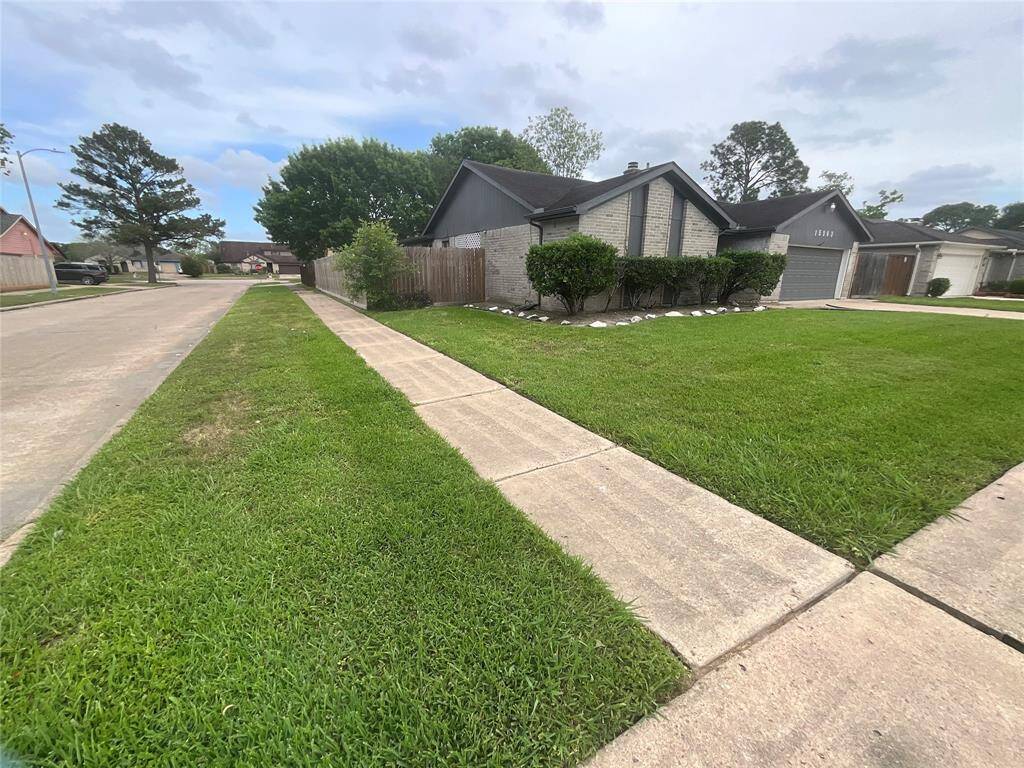This photo shows a charming corner-lot home with a well-maintained front yard, lush green grass, and neatly trimmed bushes. The house features a brick exterior and a paved driveway, situated in a quiet suburban neighborhood.