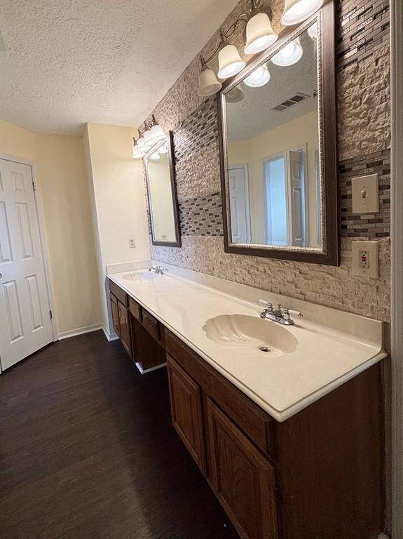 e master bathroom features a double vanity with shell-shaped sinks, framed mirrors, and stylish wall tile accents. It includes a glass walk-in shower, two walk-in closets, and a private enclosed toilet area. Dark wood-style flooring ties the space together with a warm, cohesive look.