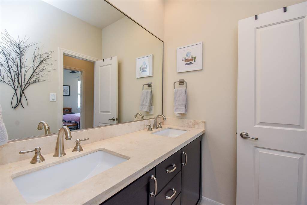 This bathroom features dual sinks, shining delicately framed wall-wide mirror and easy access to bedrooms.