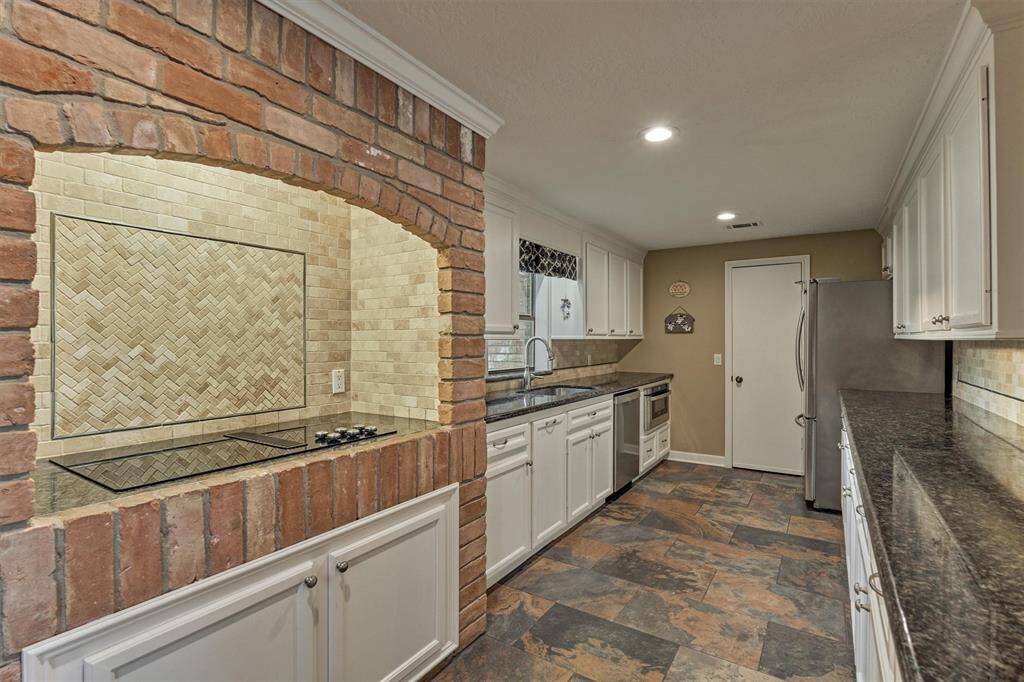 The kitchen features a unique brick archway over the cooktop, complemented by a herringbone tile backsplash. Sleek granite countertops, white cabinetry, and stainless steel appliances complete the space, while slate-look tile flooring adds warmth and character.