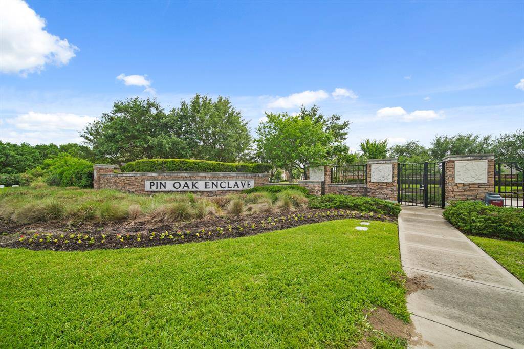 The photo shows the entrance to a residential community, "Pin Oak Enclave," featuring a well-maintained landscaped area with lush greenery, ornamental grasses, and colorful flowers. The area conveys a welcoming and serene neighborhood atmosphere.