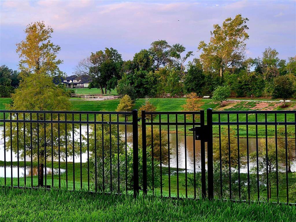 This photo showcases a serene backyard view with a black metal fence overlooking a lush green landscape, a small pond, walking path and distant trees, creating a peaceful and picturesque setting.