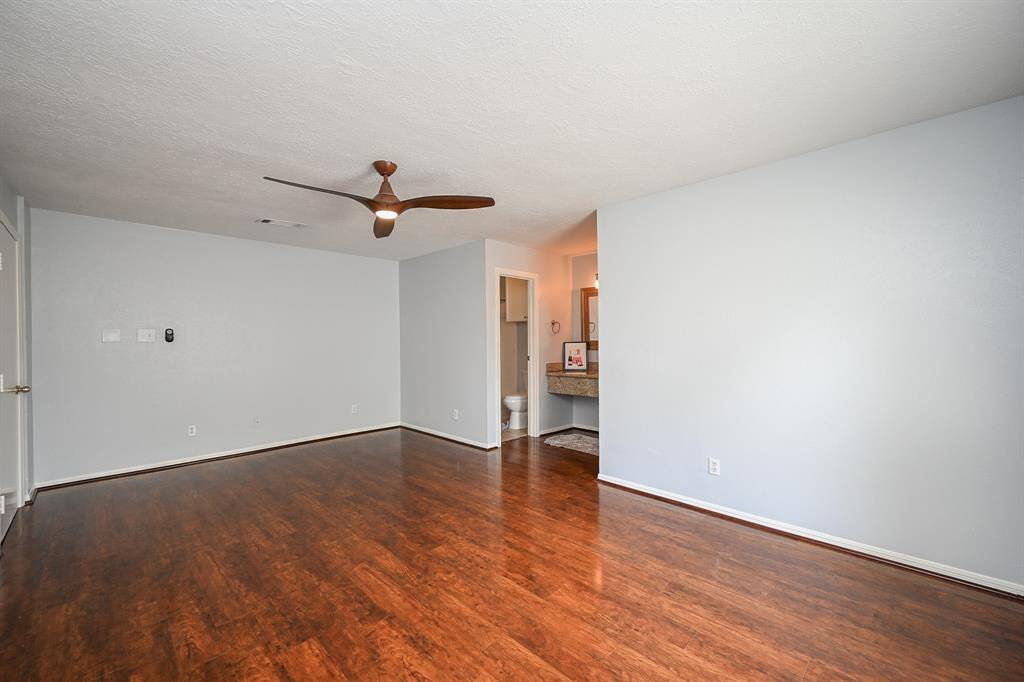 Spacious room with polished wood floors, light gray walls, and a modern ceiling fan. Features an open doorway leading to a bathroom area with a vanity mirror. Clean and bright, ready for personalization.