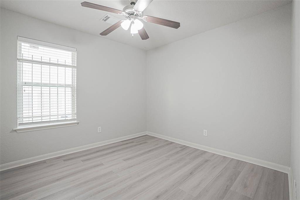 This room features light gray walls and flooring, a large window with blinds for natural light, and a ceiling fan for comfort. Ideal for a bedroom or office.