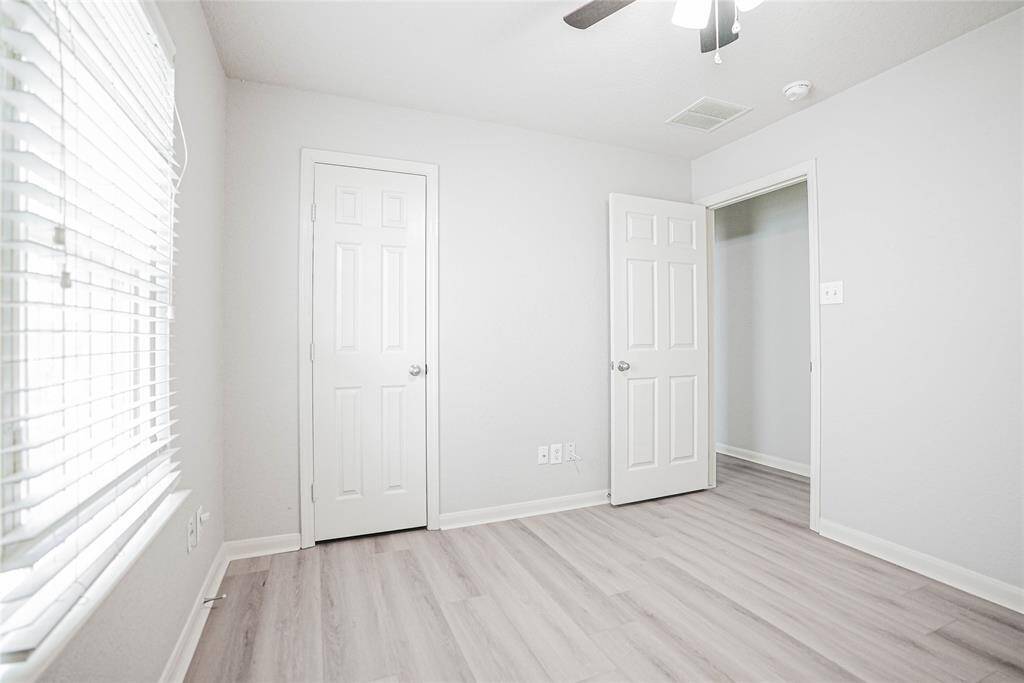 Bright, minimalist bedroom with light wood flooring, two white doors, and a window with blinds. Ceiling fan provides comfort and modern touch. Perfect for a cozy retreat.
