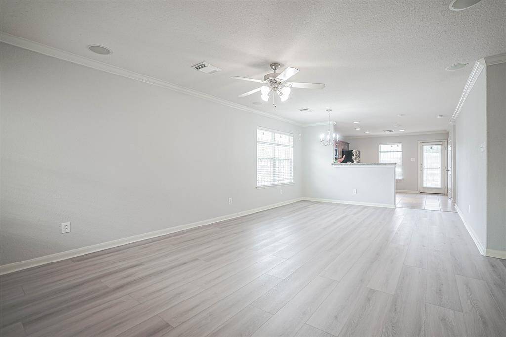 This photo displays a spacious, open-concept living area with light wood flooring, neutral walls, and abundant natural light from large windows. The space includes a ceiling fan and a view into the kitchen, which features a breakfast bar and stylish lighting fixtures.